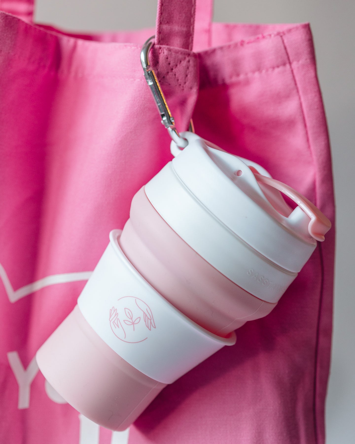 A pink and white silicone reusable coffee cup is clipped with a carabiner to a pink cloth tote bag