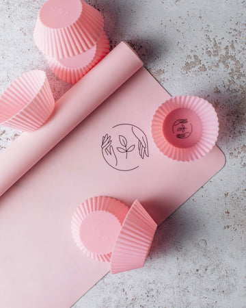 Partially rolled up pink silicone reusable baking mat featuring the Sasstainable logo on a marble surface, with a few pink silicone cupcake cases on top.