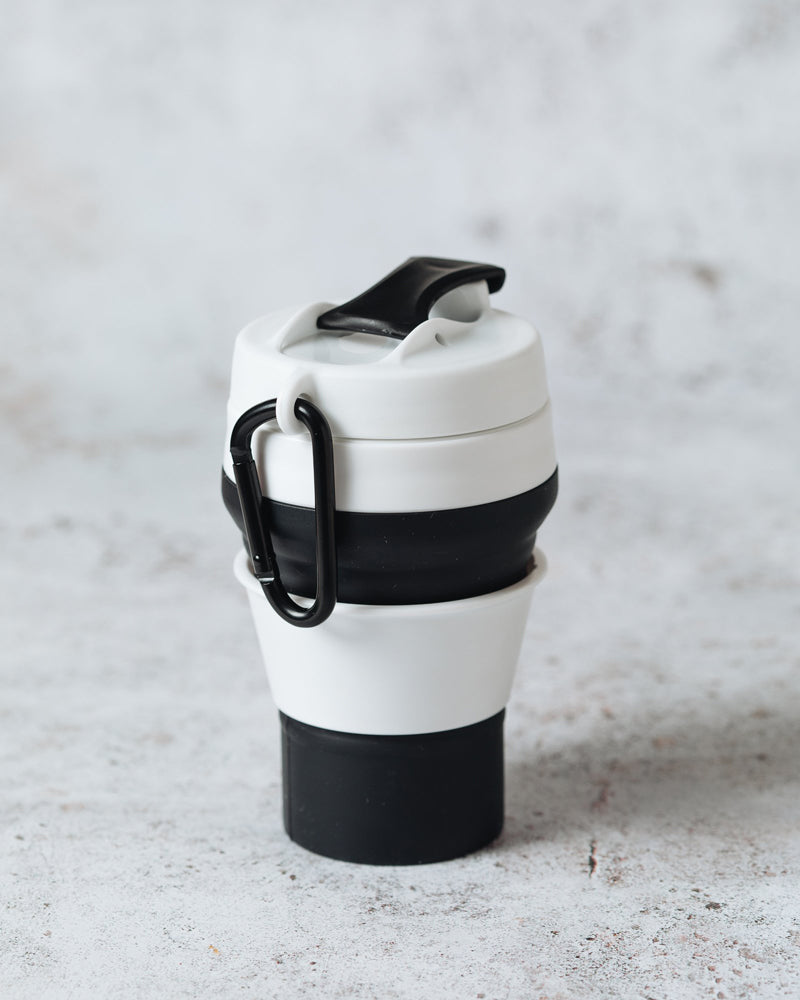 Black and white collapsible cup expanded and sitting upright on a marble surface, with a view of the carabiner clip.