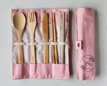 Bamboo spoon, fork, knife, chopsticks, straw cleaner and straw in a pink canvas case, next to a rolled up canvas vase, on a white background.