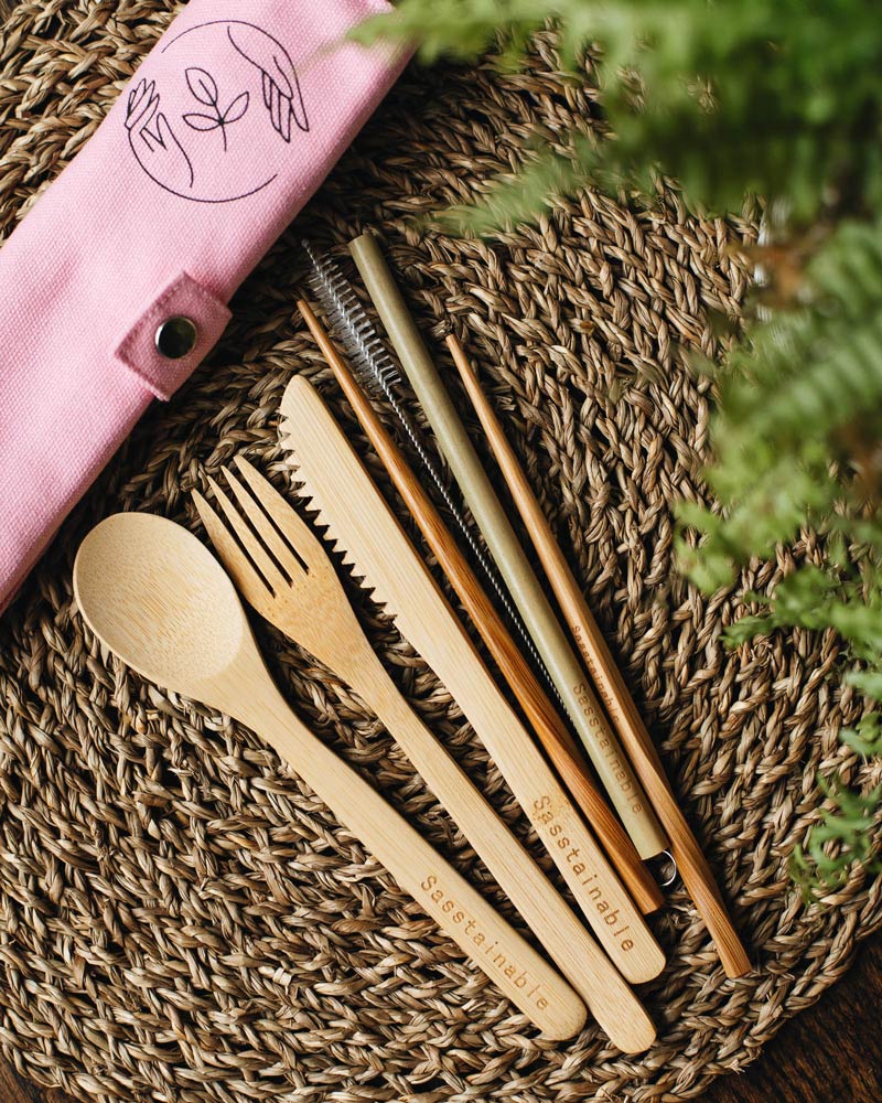 Bamboo spoon, fork, knife, chopsticks, straw and cleaner on a straw placemat with a pink rolled up canvas case with leaves in the foreground.
