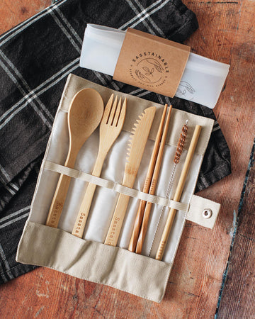 Folded up silicone pouch with a cardboard band around it, and open beige canvas case containing bamboo spoon, fork, knife, chopsticks, straw cleaner and straw on top of a checked cloth.