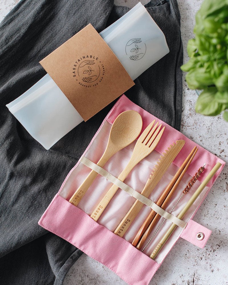 Folded up silicone pouch with a cardboard band around it, and open pink canvas case containing bamboo spoon, fork, knife, chopsticks, straw cleaner and straw on top of a dark cloth.