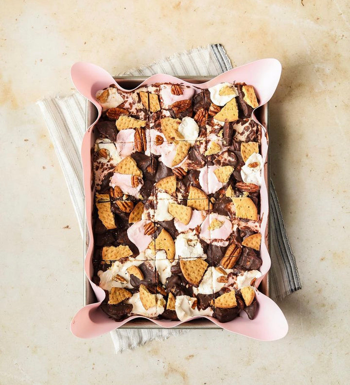 Aerial shot of tin baking pan lined with pink reusable baking sheet containing s'mores slice, sat on a tea towel