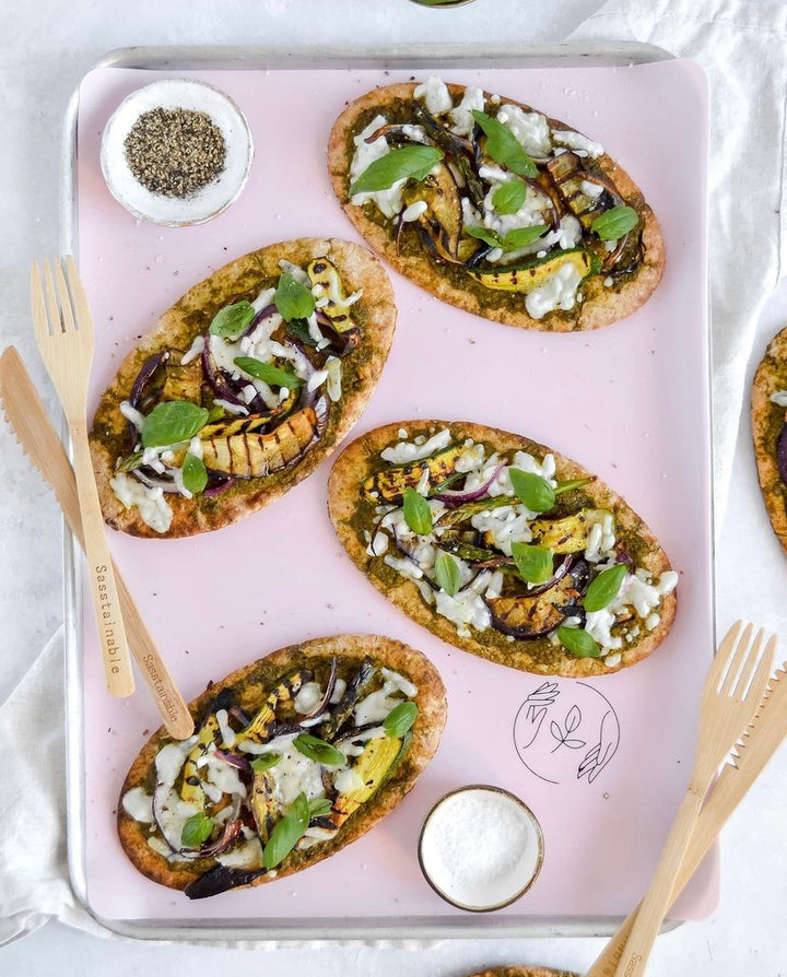 Flatbreads topped with pesto and vegetables on a pink silicone baking mat surrounded by bamboo cutlery
