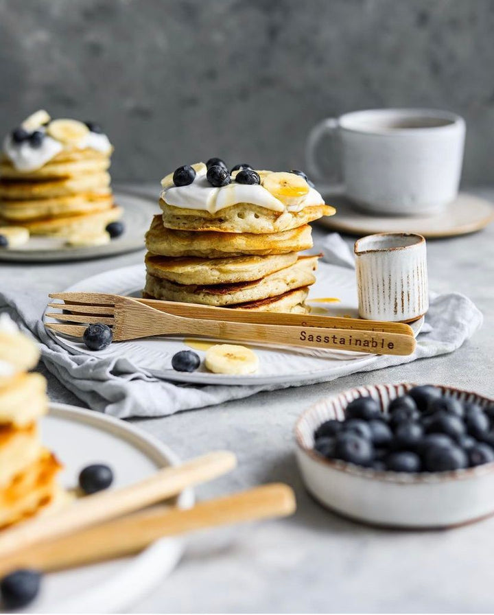 stack of pancakes surrounded by blueberries and bamboo cutlery