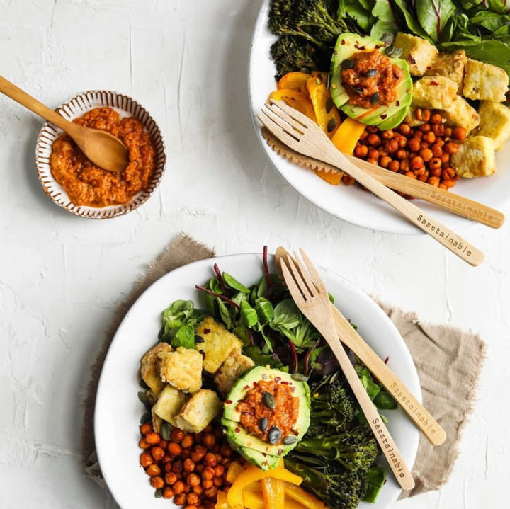 Bowl of food including avocado, peppers, chickpeas and greens with bamboo cutlery.