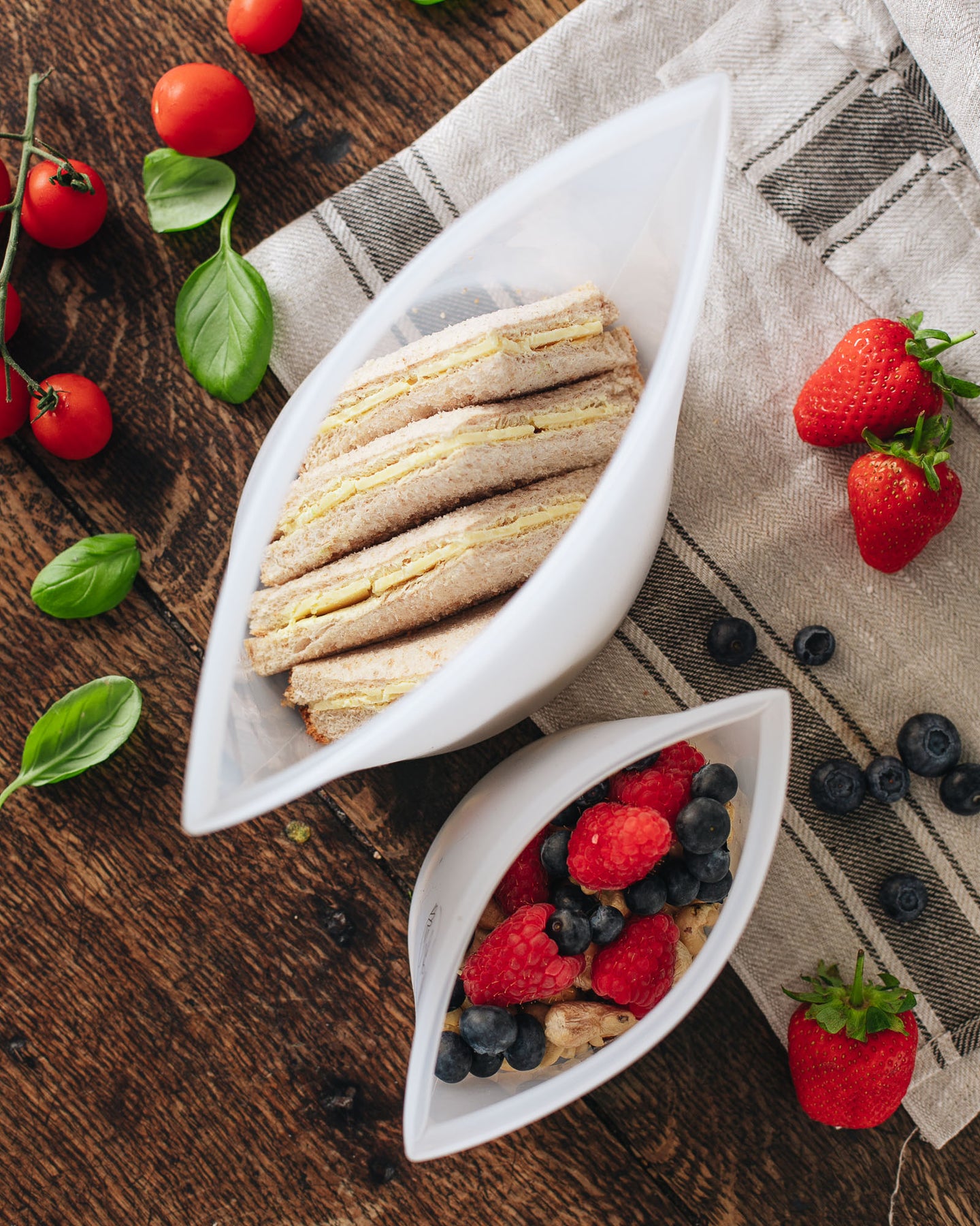Aerial view of 2 open silicone pouches; the large contains a sandwich and the small contains berries. They are sitting on a tea towel on a wooden surface with berries and tomatoes lying next to them.