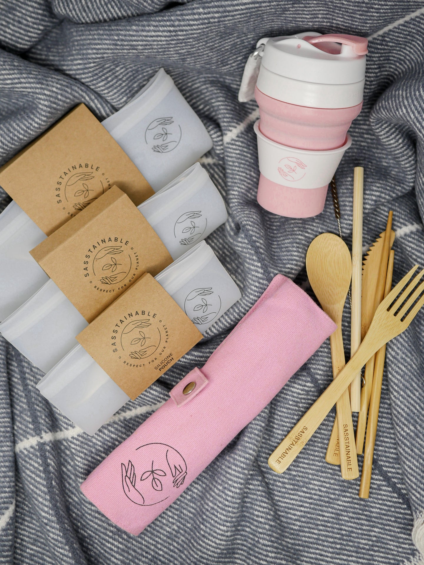 Three silicone pouches, a bamboo cutlery set, a pink cutlery case and pink silicone reusable cup on a blue cloth
