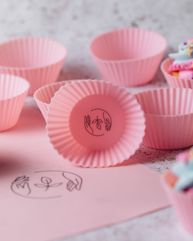 Pink silicone reusable cupcake cases featuring a black Sasstainable logo atop a pink reusable baking sheet with some iced cupcakes in the background, on a marbled surface.