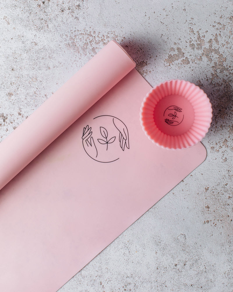 Partially rolled up pink silicone reusable baking mat featuring the Sasstainable logo on a marble surface, with a stack of pink silicone cupcake cases on top.