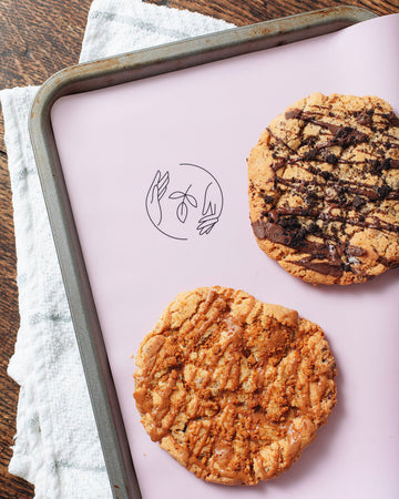 Two large cookies atop a pink silicone reusable baking mat, lining a metal tray sat on a tea towel on a wooden surface. 