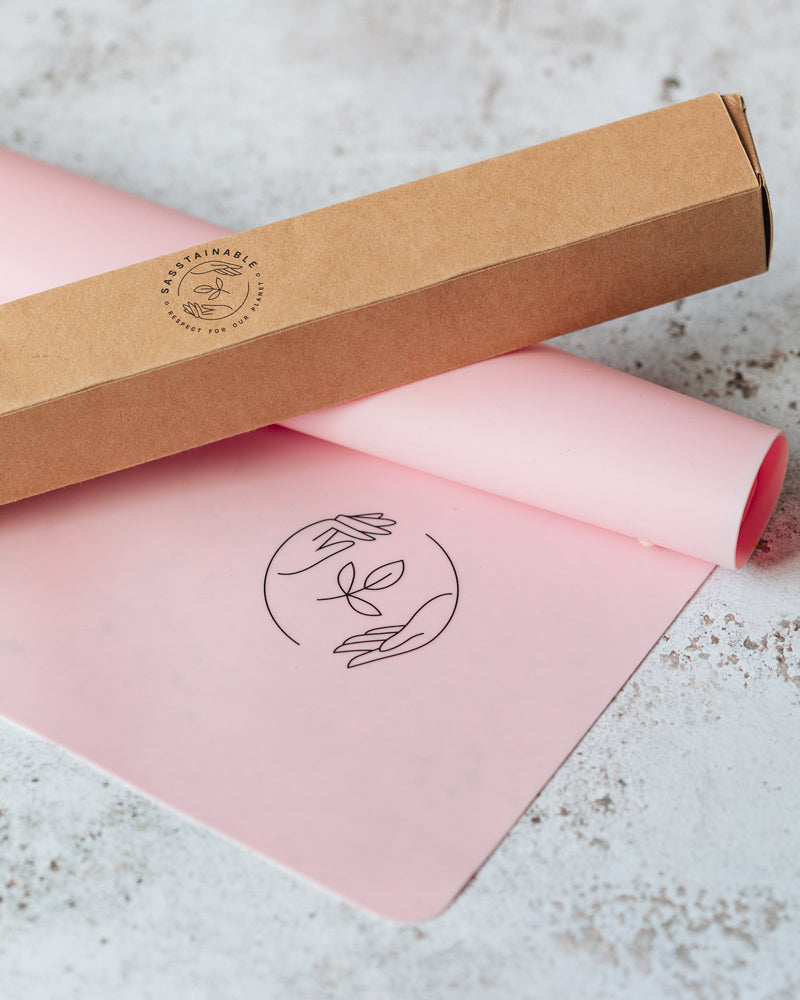 A partially rolled pink silicone reusable baking mat on a marbled surface, with a branded brown cardboard box on top of it.