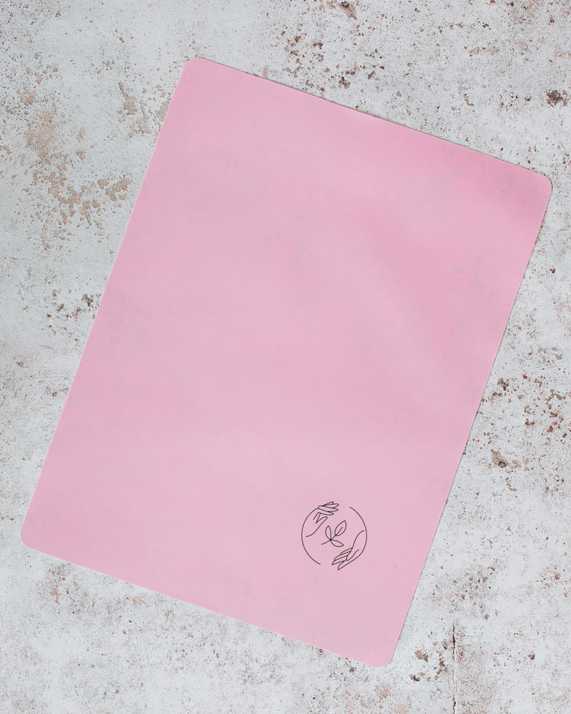 A pink silicone reusable baking mat featuring the black Sasstainble logo, sat on a marbled surface.