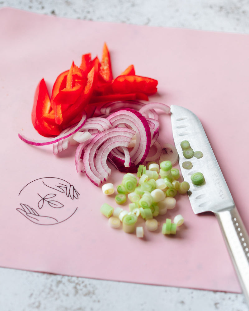 A large chopping knife sits aside of chopped spring onions, sliced red onion and sliced red pepper on top of a pink silicone reusable baking mat, on a white marble surface.