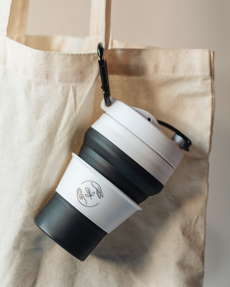 Black and white collapsible silicone cup with lid, clipped to a tote bag with a carabiner.