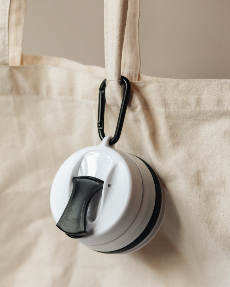 Black and white silicone cup in its collapsed state, clipped to a tote bag with a carabiner.