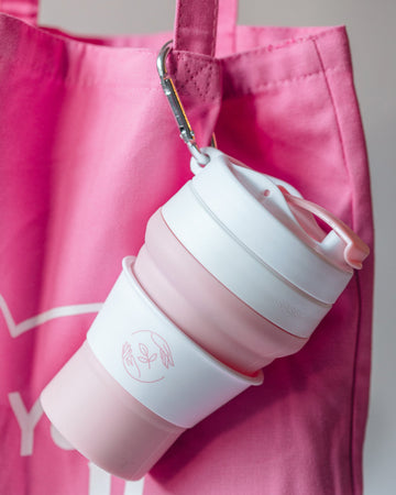 Pink and white collapsible silicone cup with lid, clipped to a tote bag with a carabiner.