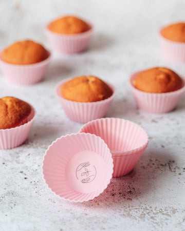 An empty pink silicone reusable cupcake case with a black Sasstainable logo next to a stack of cases, with un-iced muffins in pink cases in the background on a marble surface.