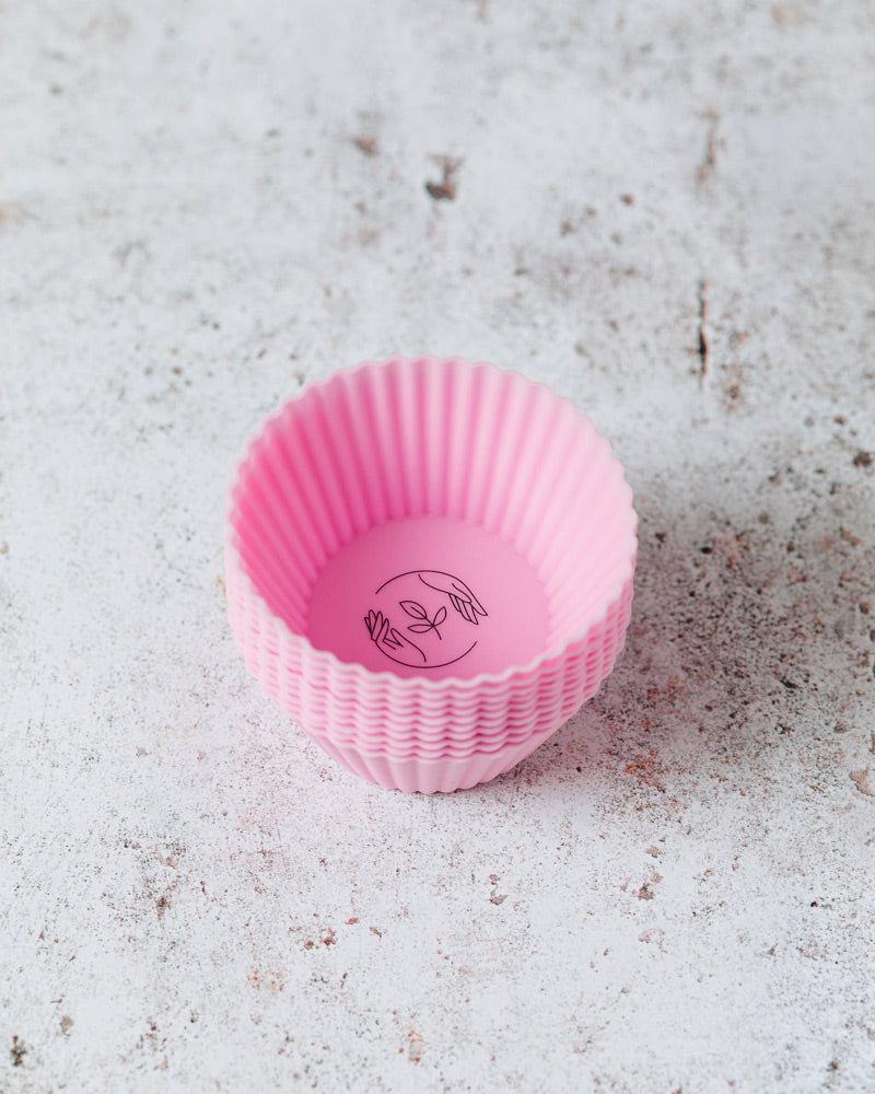 A stack of pink reusable silicone cupcake cases featuring the black Sasstainable logo atop of a marbled surface. 