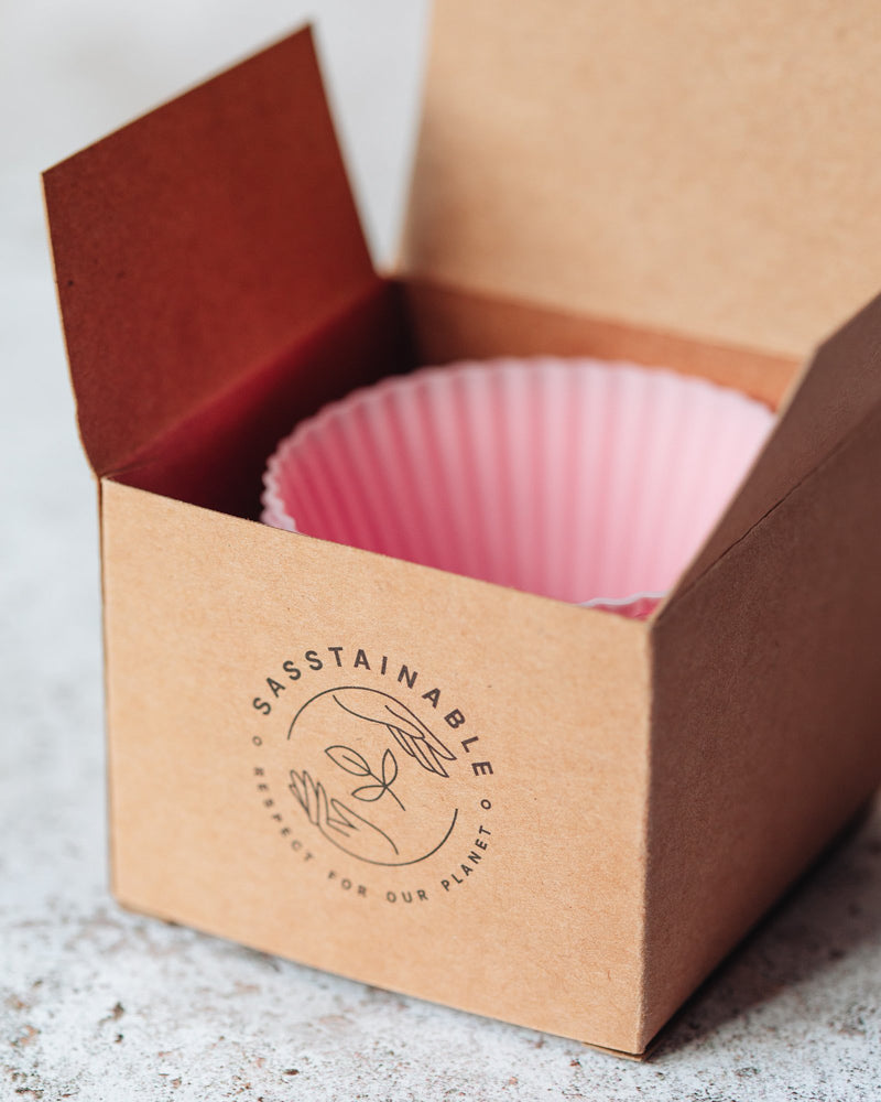 A stack of pink reusable silicone cupcake cases inside of an open brown cardboard box with the Sasstainable logo on the front.