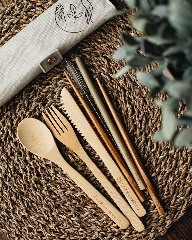 Bamboo spoon, fork, knife, chopsticks, straw and cleaner on a straw placemat with a beige rolled up canvas case with leaves in the foreground. 