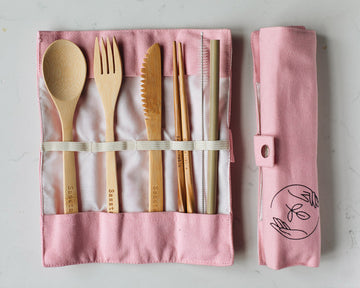 Bamboo spoon, fork, knife, chopsticks, straw cleaner and straw in a pink canvas case, next to a rolled up canvas vase, on a white background.