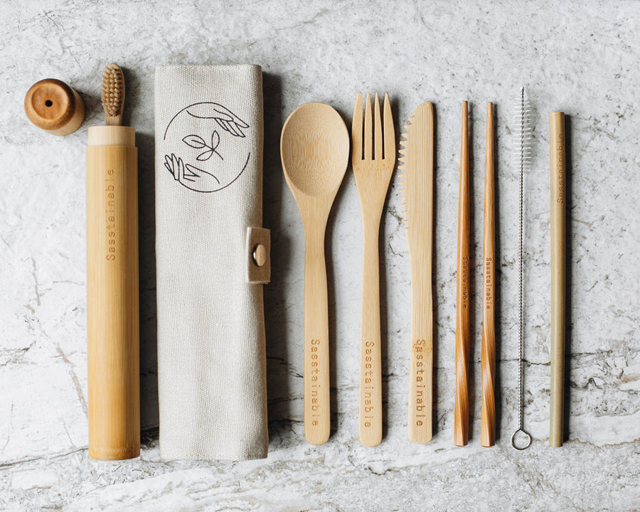 From left to right: bamboo toothbrush peeking out of bamboo tube with lid off, a beige canvas roll buttoned up with the Sasstainable logo, a bamboo spoon, fork, knife, chopsticks, straw cleaner and straw
