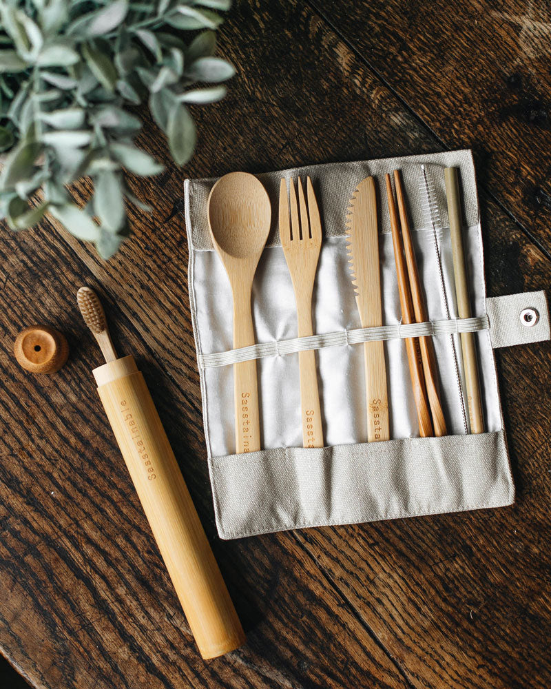 Bamboo toothbrush peeking out of bamboo tube, with bamboo spoon, fork, knife, chopsticks, straw and cleaner tucked into elastic loops of the unrolled beige canvas case, laid flat on a wooden table with some leaves visible.