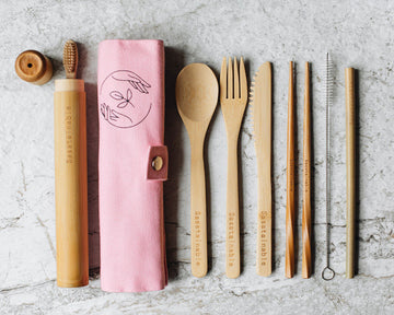 From left to right: bamboo toothbrush peeking out of bamboo tube with lid off, a pink canvas roll buttoned up with the Sasstainable logo, a bamboo spoon, fork, knife, chopsticks, straw cleaner and straw