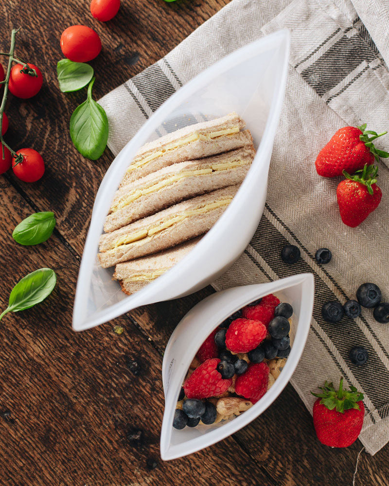 Aerial view of large silicone pouch containing a sandwich, and small silicone pouch containing berries, on a cloth-lined wooden table.