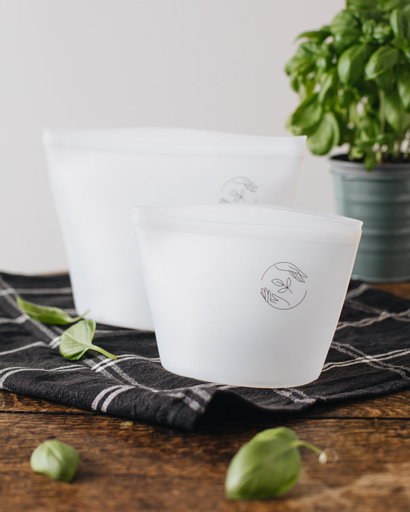 Empty large and small pouches standing side by side on a table, topped with a checked black tea towel, and a basil plant in the background.