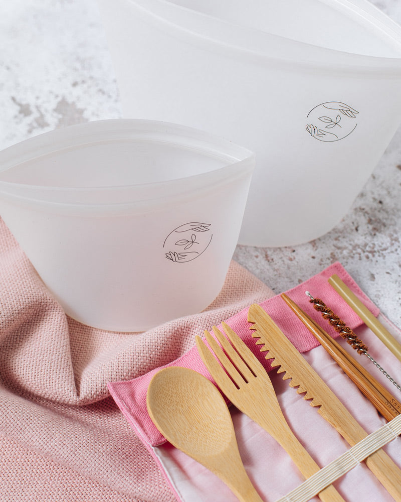 Empty large and small pouches standing side by side on a table, with an opened pink canvas case containing bamboo spoon, fork, knife, chopsticks, straw and cleaner.