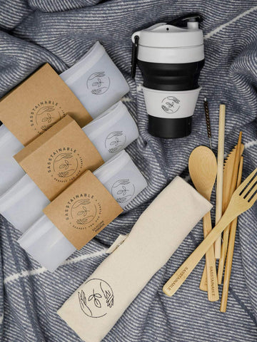 Picnic bundle: natural bamboo cutlery set, collapsible black silicone coffee cup, reusable silicone pouches in three sizes pictured on a blanket