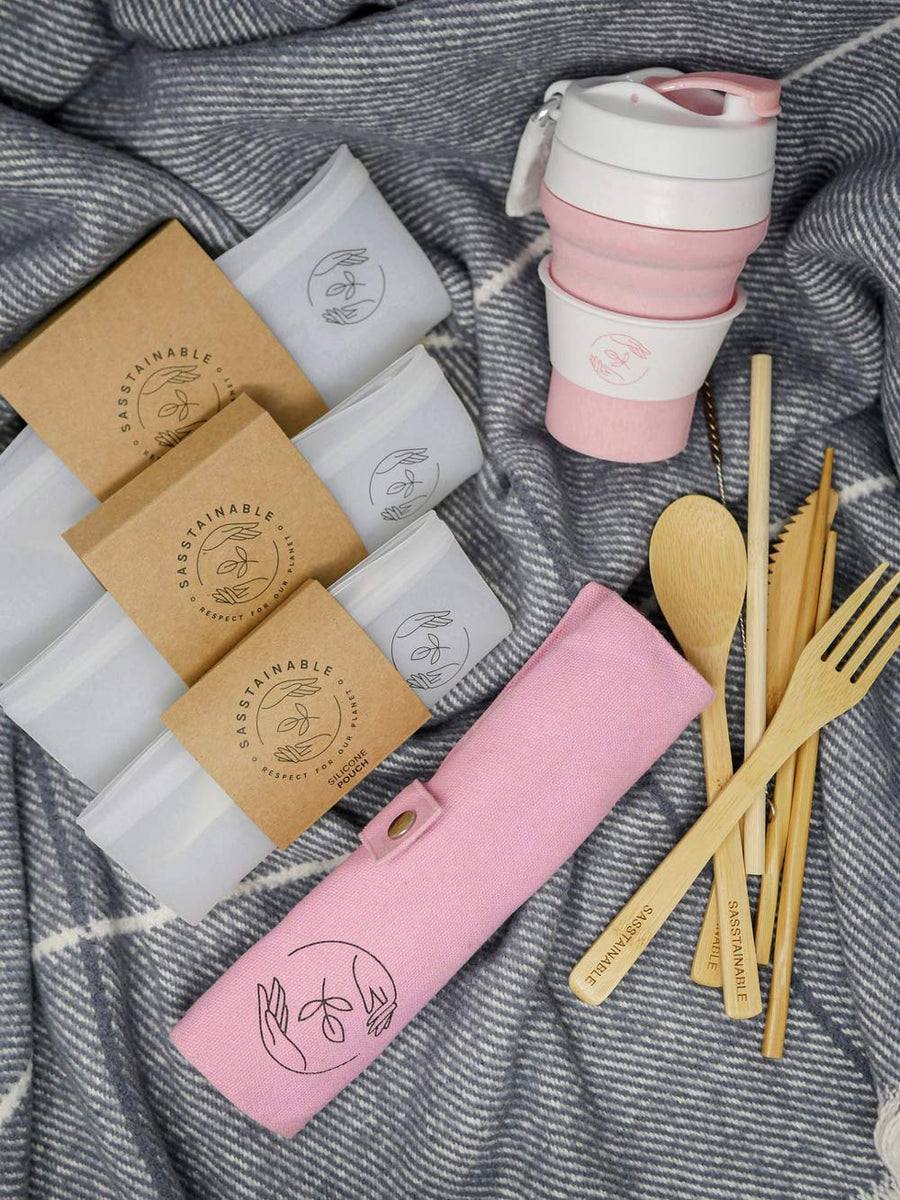 Picnic bundle: pink bamboo cutlery set, collapsible pink silicone coffee cup, reusable silicone pouches in three sizes pictured on a blanket