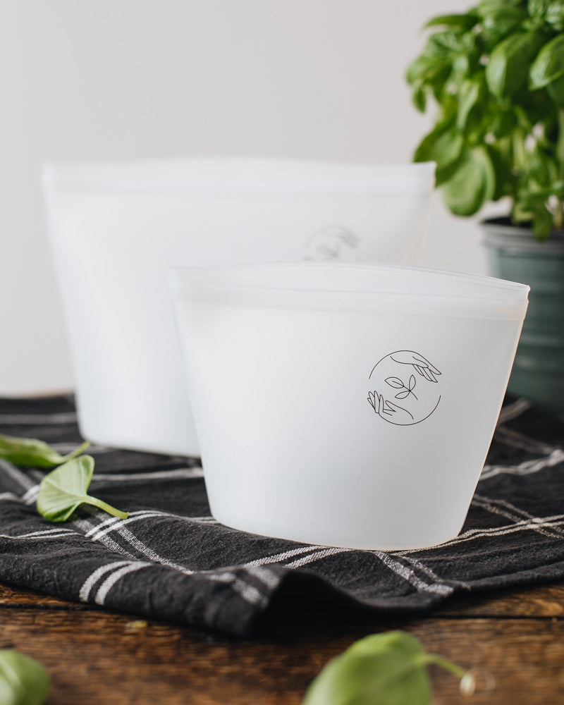 Empty small and large silicone pouches on a checked cloth with a basil plant in the background.