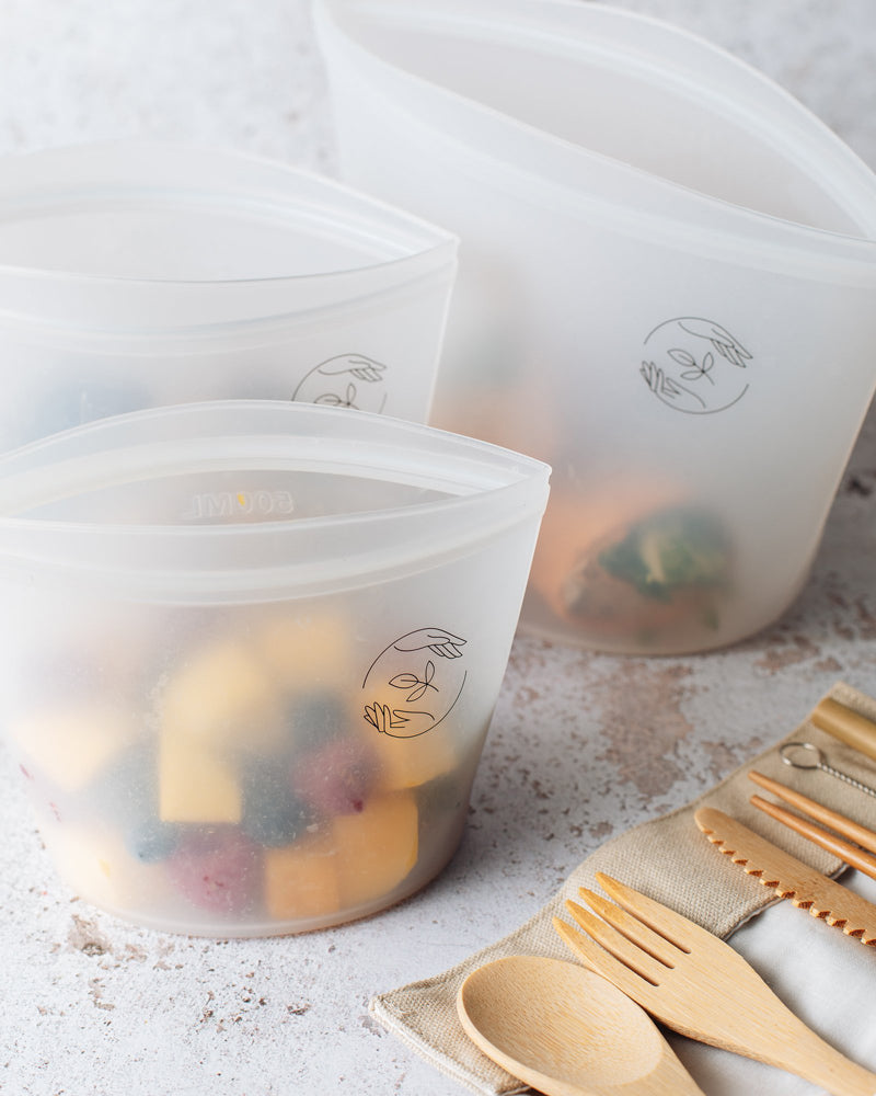 Small silicone pouch filled with fruit, in front of medium and large reusable pouches, with bamboo cutlery in the foreground.