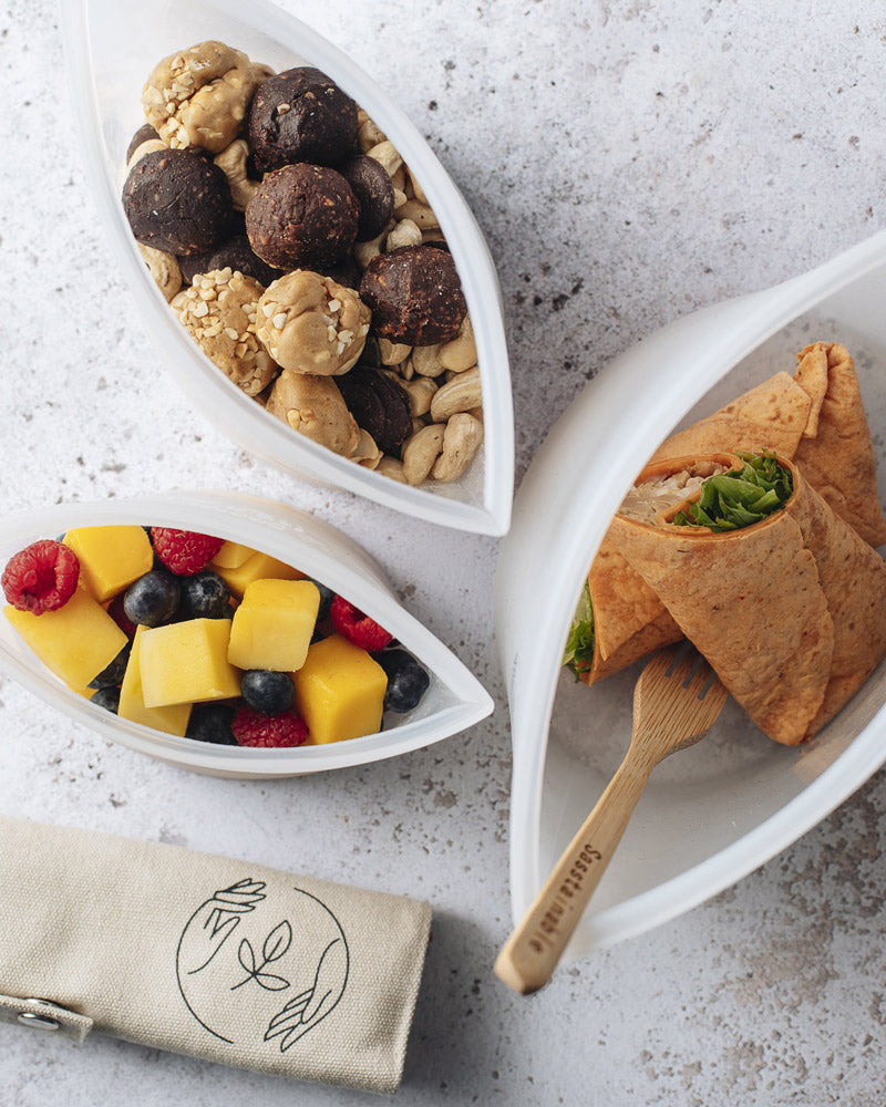 Aerial view of large silicone pouch containing a salad wrap, medium pouch containing nuts and protein balls, and a small silicone pouch containing cubed mango, blueberries and raspberries on a marble surface, with a canvas cutlery roll nearby, and a bamboo fork in the pouch with the sandwich.