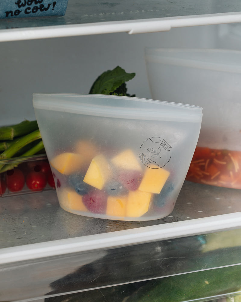 A small silicone reusable pouch containing cubed mango, raspberries and blueberries sat on an open fridge shelf with vegetables in the background.