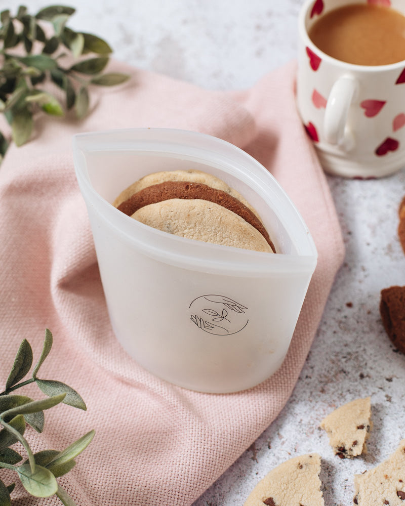 Small silicone pouch containing 3 large cookies on a pink cloth, atop a marbled surface with a mug full of milky tea in the background. 