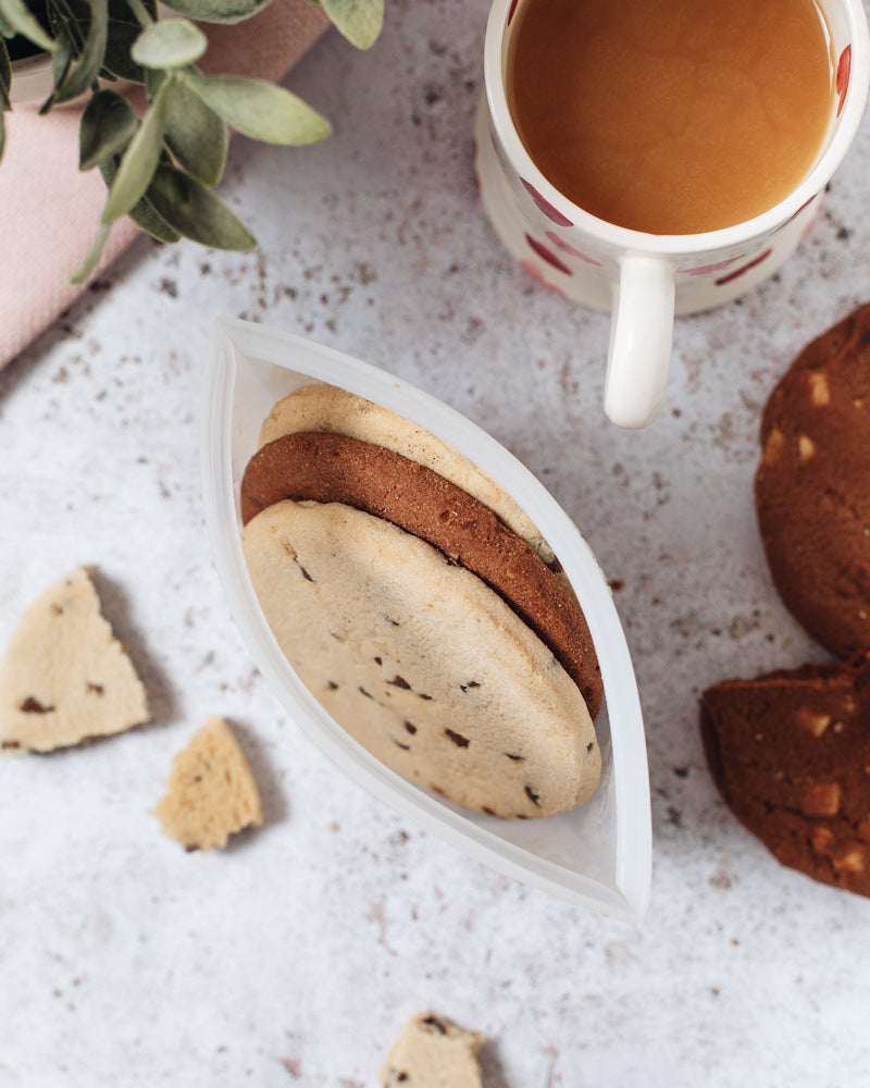 Aerial shot of a small silicone pouch containing 3 large cookies atop a marbled surface with a mug full of milky tea and broken cookies around it.