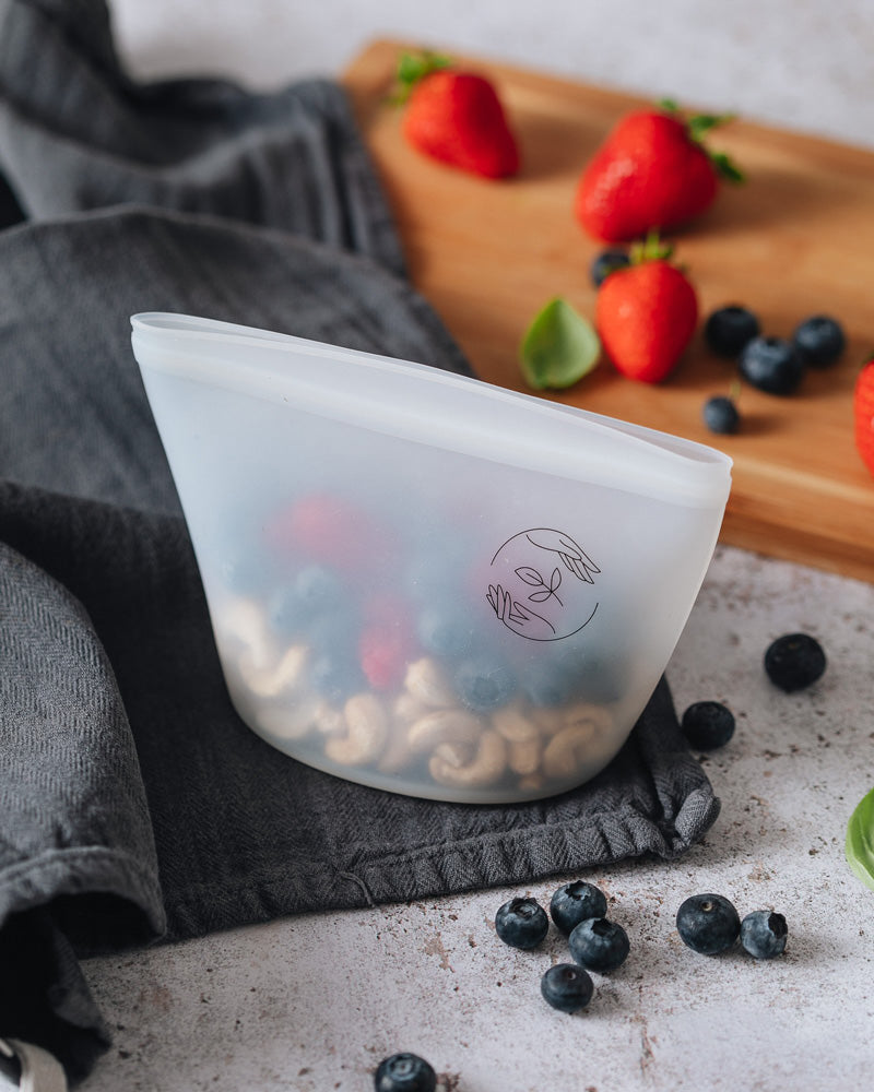 Small silicone reusable pouch containing cashew nuts and berries, on top of a dark cloth with a wooden chopping board in the background, atop a marble surface surrounded by mixed berries.