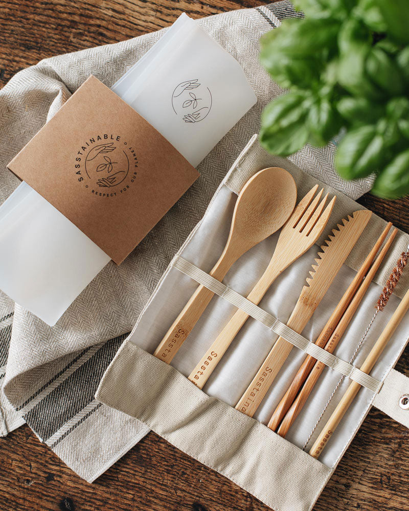 Folded up silicone pouch with a cardboard band around it, and open beige canvas case containing bamboo spoon, fork, knife, chopsticks, straw cleaner and straw on top of a natural coloured cloth atop a wooden surface.