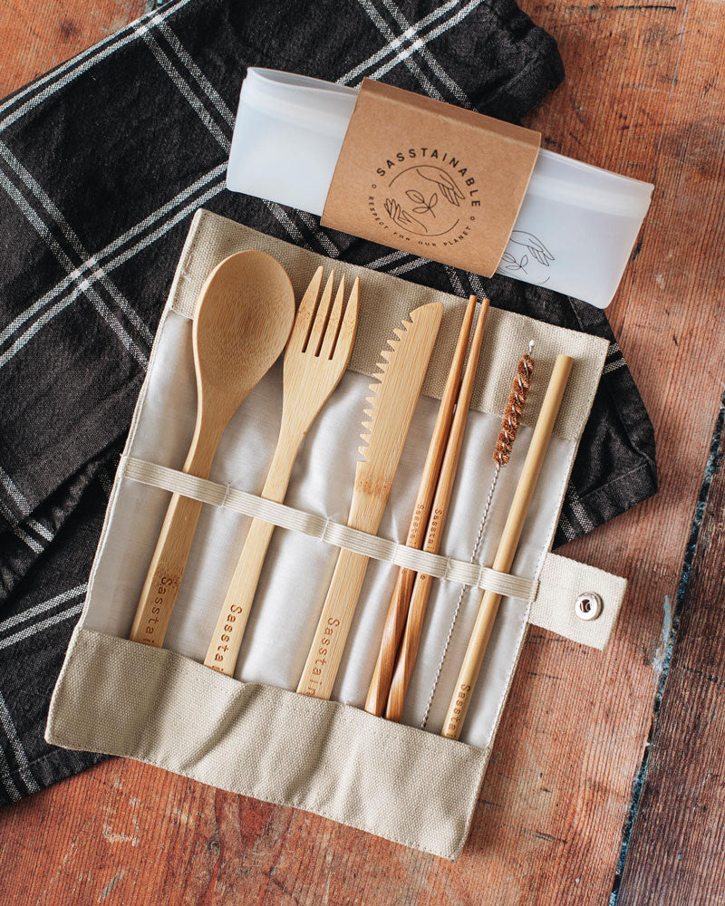 Folded up silicone pouch with a cardboard band around it, and open beige canvas case containing bamboo spoon, fork, knife, chopsticks, straw cleaner and straw on top of a checked cloth.