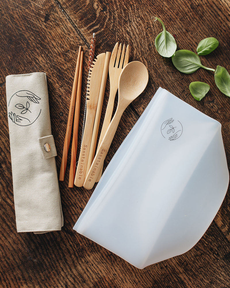 Bamboo cutlery, silicone pouch and beige canvas case with Sasstainable logo on a wooden surface with basil leaves laying nearby.