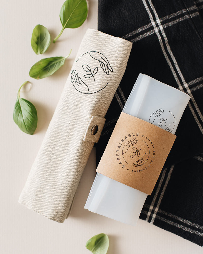 Beige canvas case rolled up, and silicone pouch folded flat lying on a checked cloth, atop a white surface, with some basil leaves strewn about.