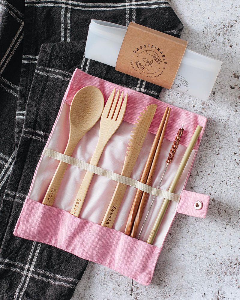 Folded up silicone pouch with a cardboard band around it, and open pink canvas case containing bamboo spoon, fork, knife, chopsticks, straw cleaner and straw on top of a checked cloth.