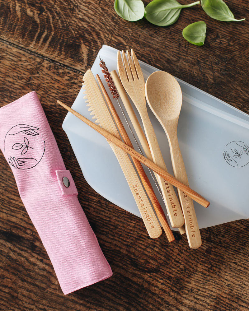 Bamboo cutlery, silicone pouch and pink canvas case with Sasstainable logo on a wooden surface.