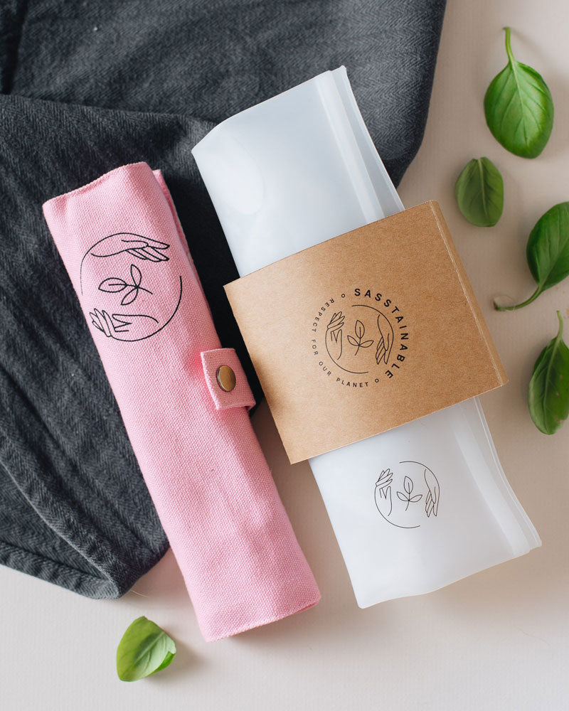 Pink canvas case rolled up, and silicone pouch folded flat lying on a dark cloth, atop a white surface, with some basil leaves strewn about.