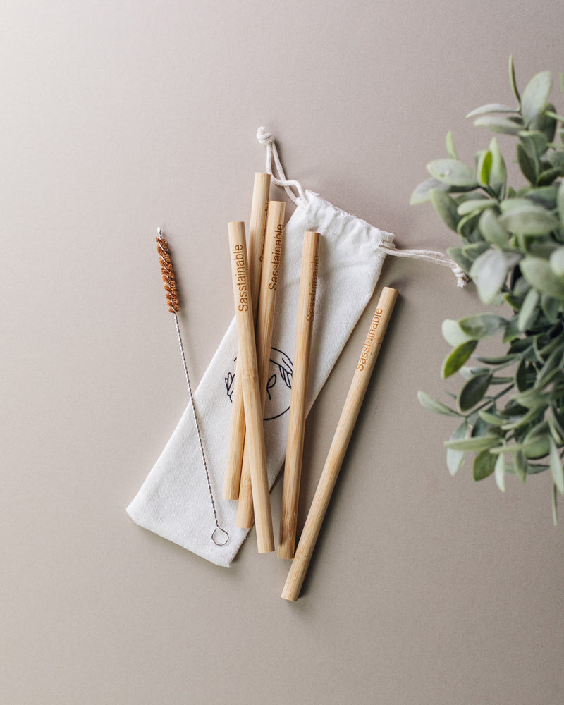 Five bamboo straws and a straw cleaner laid on a white canvas drawstring pouch featuring the Sasstainable logo on a plain surface, with leaves in the foreground.